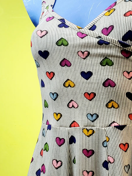wearing cotton fit and flare sleeveless dress with surplice bodice and v neckline. Dress has a full knee length skirt and pockets. Warm light beige background with delicate vertical stripe pattern and all over staggered cartoony red, yellow, light blue, royal blue, purple, and pink heart pattern. Showing torso close up