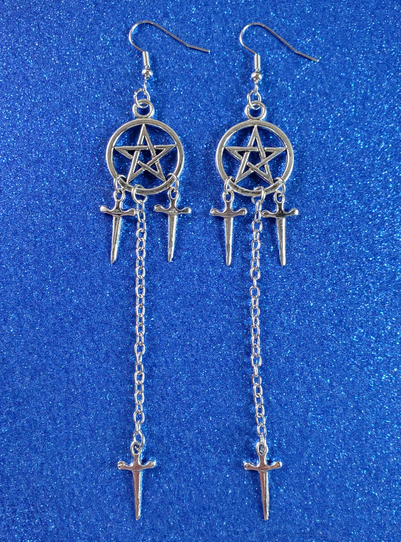 pair of pentagram dangle earrings in shiny silver metal with both a pair of 3/4” dagger charms hanging from each pentagram & a 2 1/2” link chain connecting another dagger to the bottom of the earring