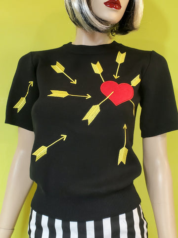black short sleeve pullover sweater features an appliqued bold red felt heart under attack from many bright yellow embroidered arrows, shown on a mannequin