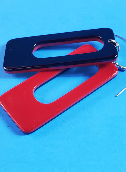 A pair of rectangular acrylic dangle earrings with rounded corners and an oblong hole in the middle. The front of the earring is deep red and the back is black