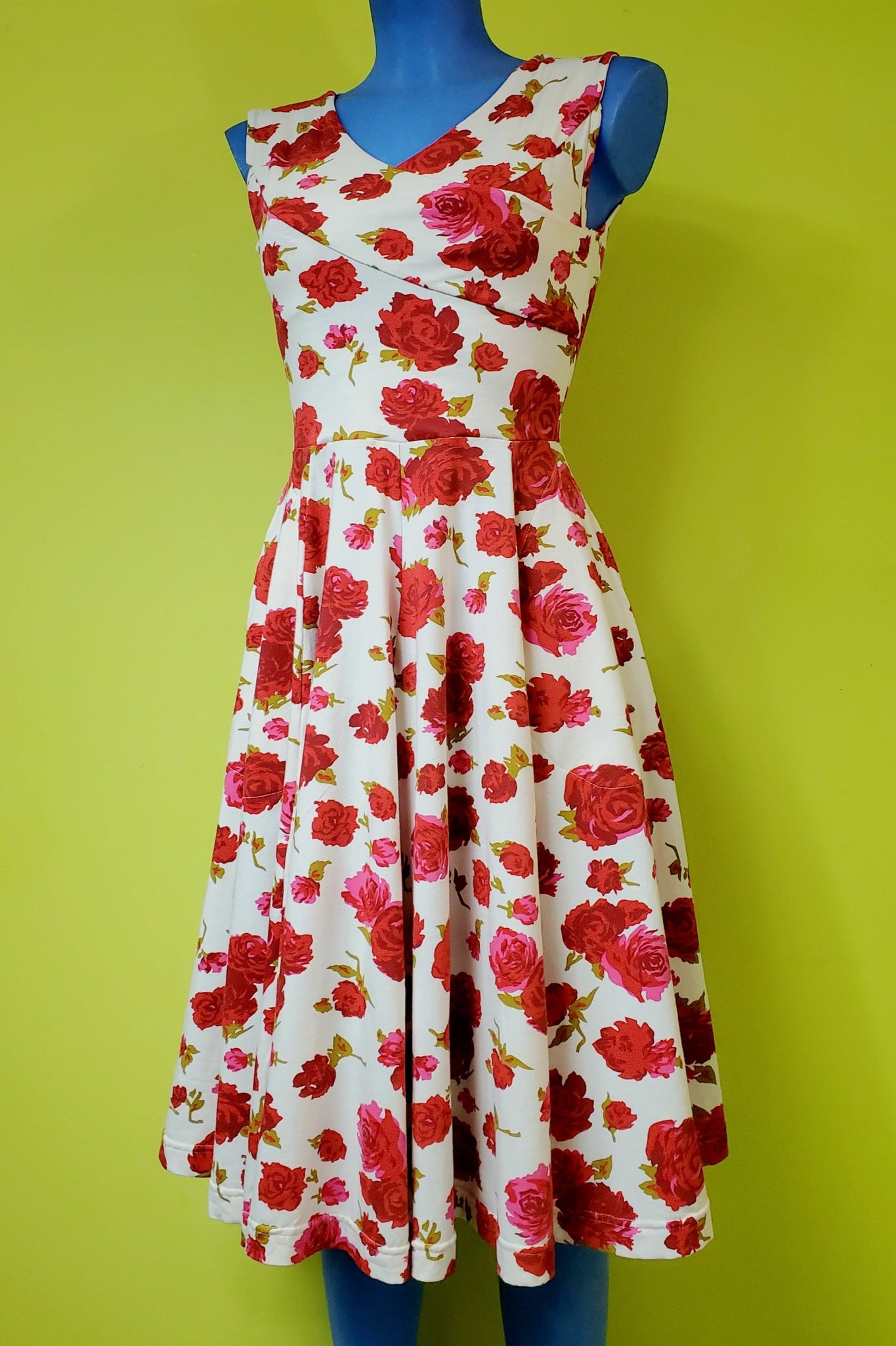 a sleeveless fit and flare dress in a stretch cotton knit creamy white background blooming pink, red, and green rose pattern, and featuring a faux-wrap surplice bodice with v-neckline, and paneled just-below-the-knee length full skirt. shown on a mannequin