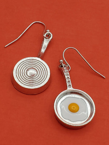 Pair of dangle earrings, each in the shape of a silver metal frying pan with white and yellow enamel sunny side up egg inside. This image shows the underside of one of the pans