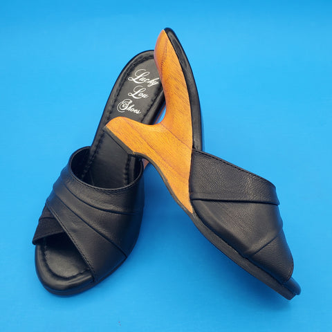 pair of wooden boomerang shaped heels with open toes and black leather & suede toe straps. The shoes have metallic gold insoles and black rubber soles. One is shown from the side to emphasize heel shape