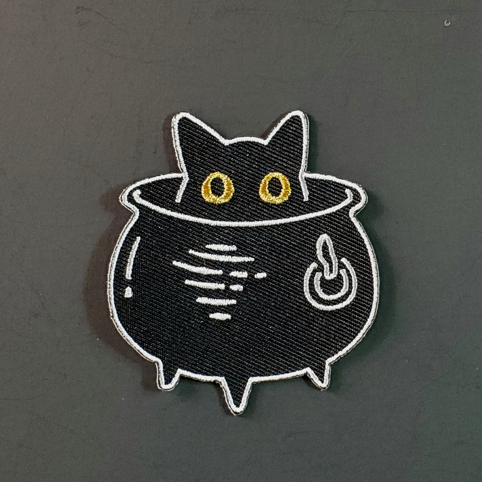 Black twill embroidered patch of a black cat with large metallic gold eyes sticking its head out of a cauldron