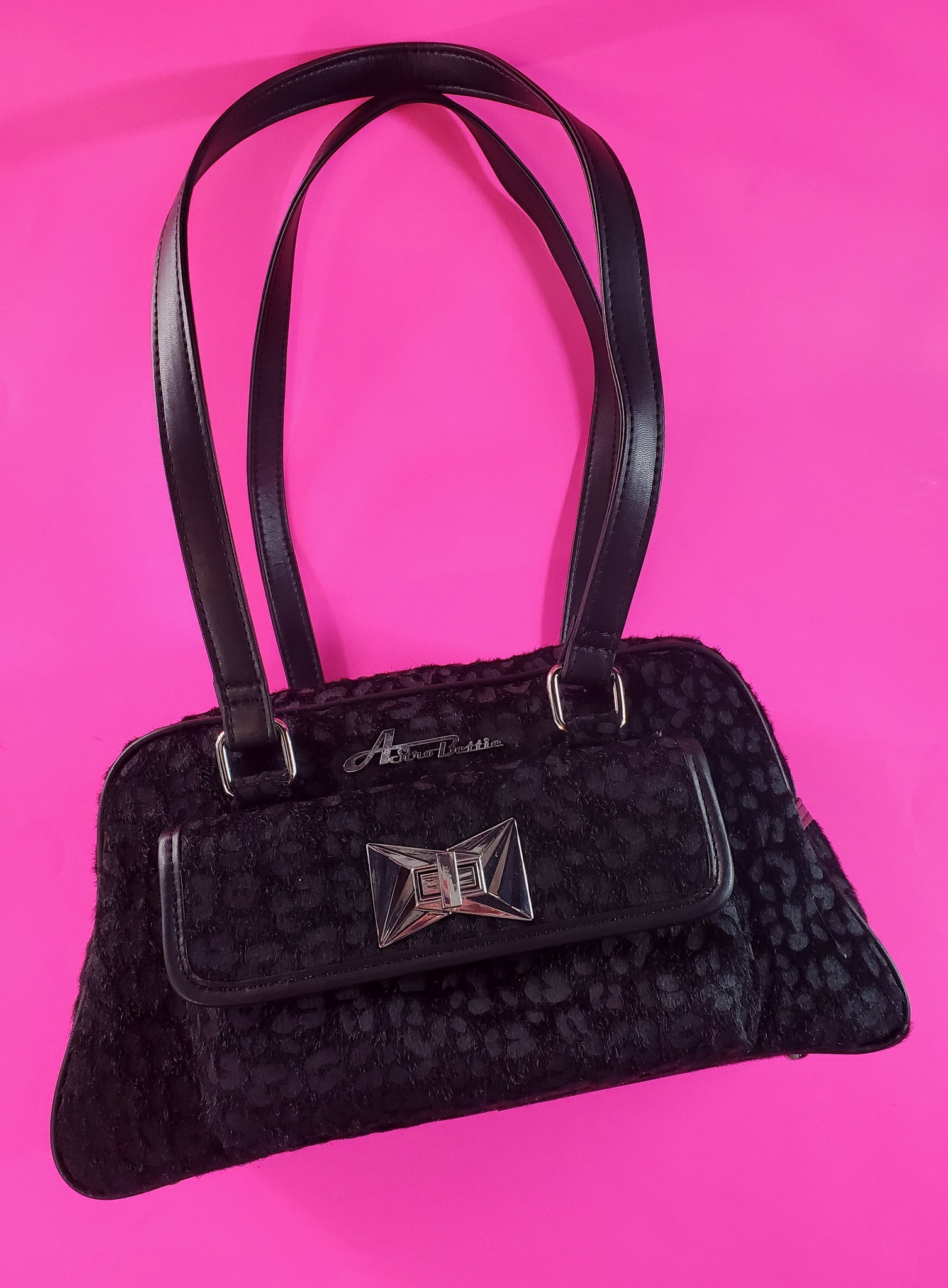 Trapezoid-shaped purse in a black on black leopard pattern printed on fuzzy faux pony with matching matte faux leather trim and shoulder straps. The large exterior pocket on the front features an Art Deco inspired silver metal clasp and the Astro Bettie logo in silver metal