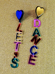 A pair of drop earrings made of mirrored acrylic letters attached to each other with jump rings spelling “LET’S” and “DANCE” in pink, green, yellow, blue, and red. Attaches to ear with heart shaped charm