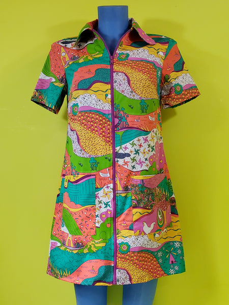 A mannequin wearing a stretch cotton mini dress with a rounded wide collar, short sleeves, and a bright purple plastic zipper running down the length of the dress. It has an intricate multicolored psychedelic pattern including pinwheels, apples, hands, birds, and other illustrations in pink, yellow, orange, blue, green, and white. Shown from front