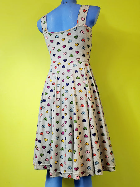 mannequin wearing cotton fit and flare sleeveless dress with surplice bodice and v neckline. Dress has a full knee length skirt and pockets. Warm light beige background with delicate vertical stripe pattern and all over staggered cartoony red, yellow, light blue, royal blue, purple, and pink heart pattern. Shown from the back