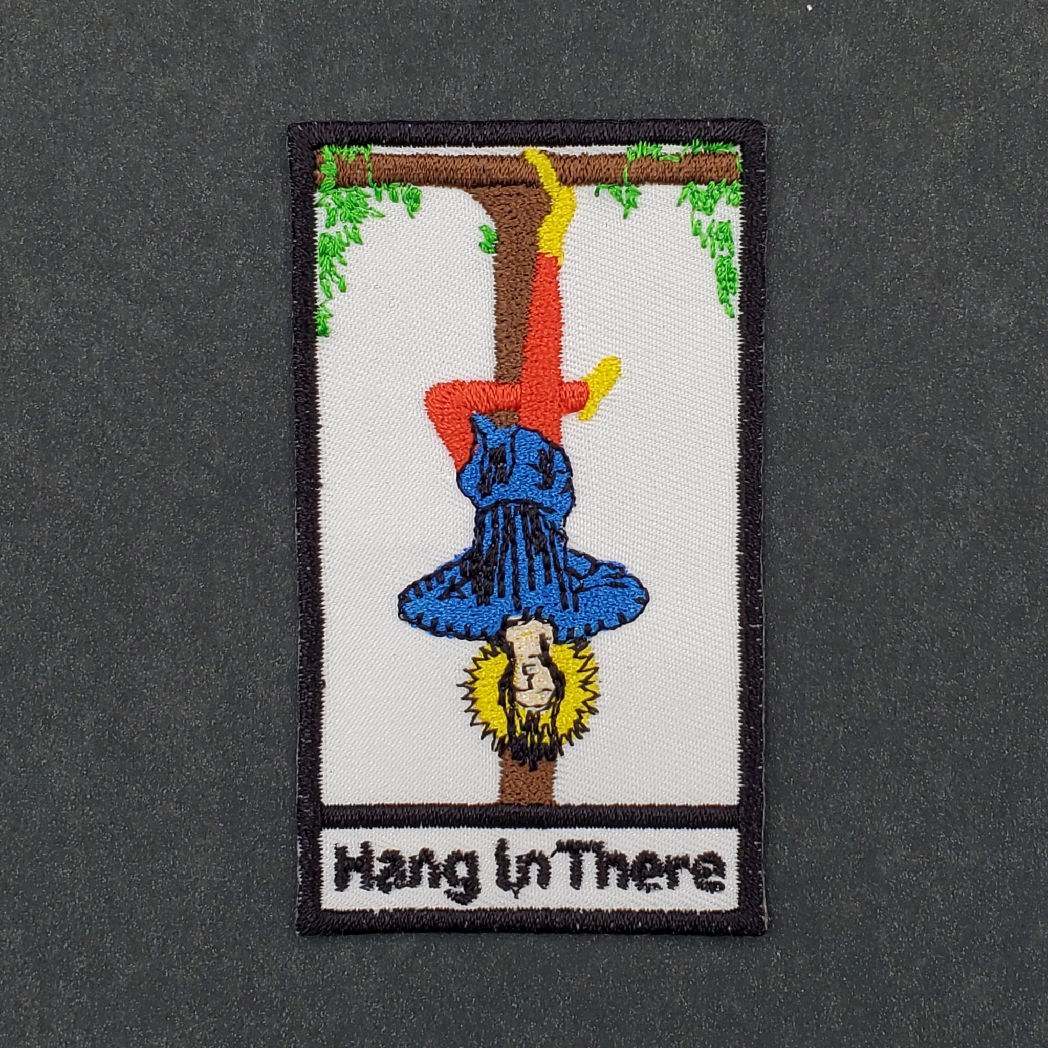 rectangular twill patch of the Hanged Man tarot card in blue, yellow, red, and black with message "Hang in there" written at bottom