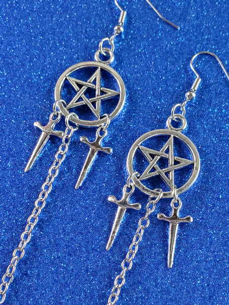 pair of pentagram dangle earrings in shiny silver metal with both a pair of 3/4” dagger charms hanging from each pentagram & a 2 1/2” link chain connecting another dagger to the bottom of the earring. showing close-up view