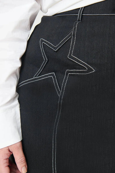 Model wearing a black denim a-line mini skirt with contrasting warm white stitching running vertically down skirt with white star embroidery on each hip and zipper/button front closure. Shown from front In close up of star embroidery detail