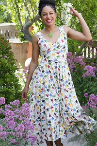 model wearing a sleeveless fit & flare stretch cotton knit dress in creamy beige background print with repeating pattern of graceful flying swallows in black, red, blue, yellow, and green, and features a faux-wrap surplice bodice with v-neckline, wide straps, and mid-calf length extra full & swing-y paneled skirt