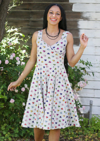 Model wearing cotton fit and flare sleeveless dress with surplice bodice and v neckline. Dress has a full knee length skirt and pockets. Warm light beige background with delicate vertical stripe pattern and all over staggered cartoony red, yellow, light blue, royal blue, purple, and pink heart pattern. Shown from the front