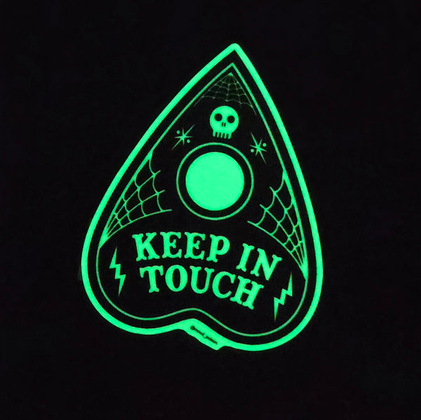 die-cut glow-in-the-dark vinyl sticker of a black Ouija planchette with illustrated spiderwebs, lightning bolts, and a miniature skull with the message to “keep in touch" shown glowing