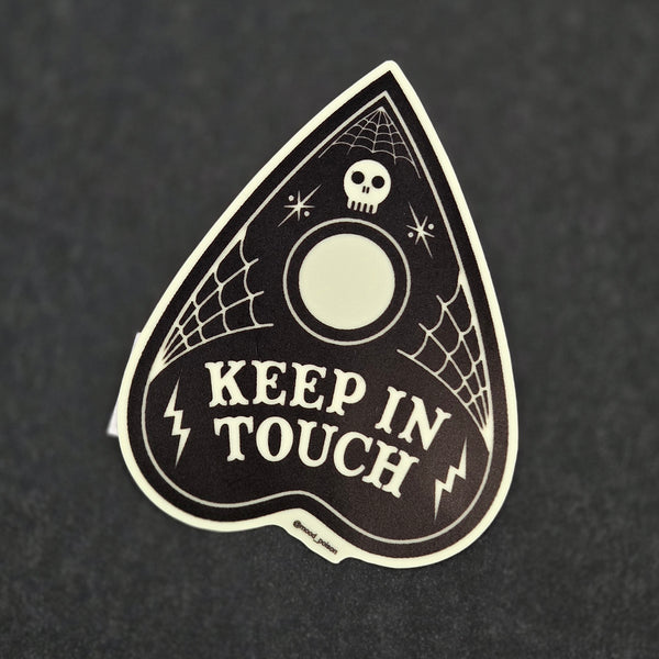 die-cut glow-in-the-dark vinyl sticker of a black Ouija planchette with illustrated spiderwebs, lightning bolts, and a miniature skull with the message to “keep in touch"