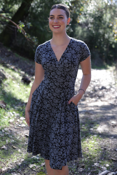 A woman wearing a stretch cotton knit short-sleeve faux wrap surplice V-neck dress with a black and lavender purple rosebud and leaf pattern with knee length full skirt. Shown standing outdoors in a wooded area.