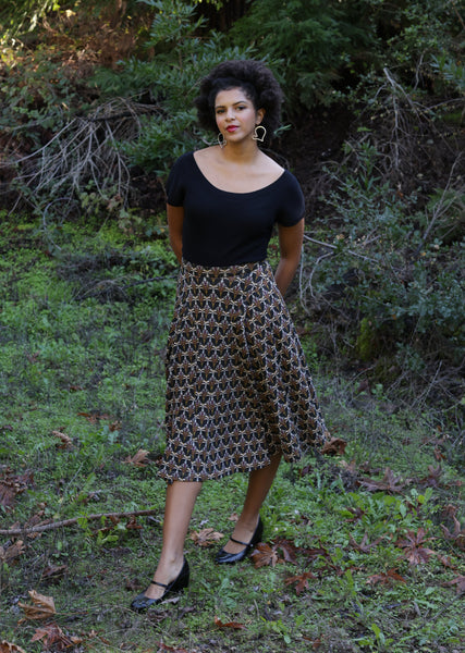 Model wearing stretch cotton knit below the knee full skirt with wide high waistband and gored detail in a cream background Art Deco style bird pattern. Shown in wooden area against trees