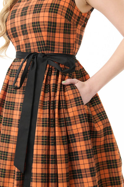 Model wearing a cotton orange, black, and yellow gold lurex plaid patterned swing dress with slash neckline, darted bodice, black cotton sash belt, and below the knee gathered skirt. Shown from three quarter angle in close up of sash belt