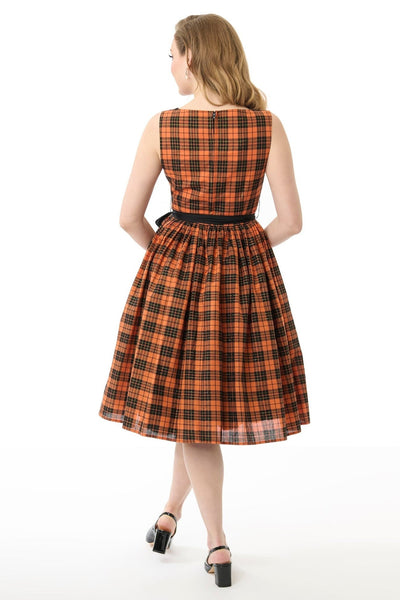 Model wearing a cotton orange, black, and yellow gold lurex plaid patterned swing dress with slash neckline, darted bodice, black cotton sash belt, and below the knee gathered skirt. Shown from back