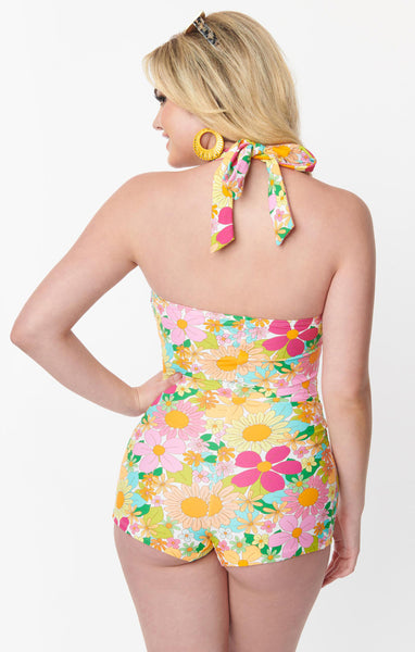 A model wearing a 50s style halter one piece. It is in a bright pink, orange, yellow, and green 70s style floral pattern. It has wide matching halter straps, a keyhole and tie detail at the bust, slightly ruched waist, and boy short style bottom. Seen from behind to show halter tie detail 