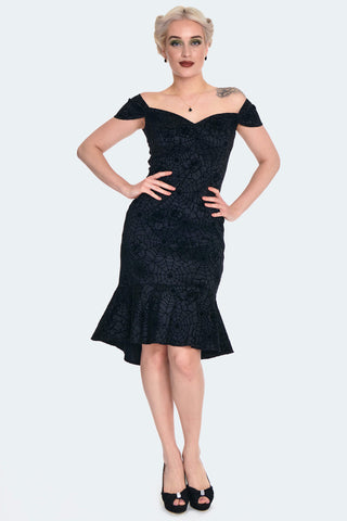 A model wearing a black wiggle dress in a flocked rose and spiderweb taffeta fabric. It has a sweetheart neckline and off the shoulder cap sleeves with an open upper back. The wiggle skirt has a high-low mermaid style hemline that ends just at the knee. Shown from the front