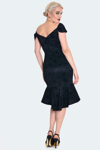 A model wearing a black wiggle dress in a flocked rose and spiderweb taffeta fabric. It has a sweetheart neckline and off the shoulder cap sleeves with an open upper back. The wiggle skirt has a high-low mermaid style hemline that ends just at the knee. Shown from the back
