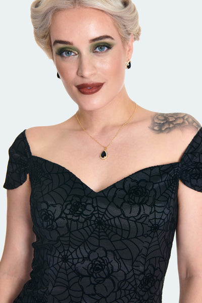 A model wearing a black wiggle dress in a flocked rose and spiderweb taffeta fabric. It has a sweetheart neckline and off the shoulder cap sleeves with an open upper back. The wiggle skirt has a high-low mermaid style hemline that ends just at the knee. Shown from the front in close up