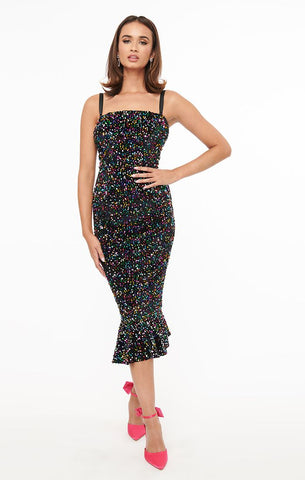 A model wearing a black velvet wiggle dress covered in multicolored rainbow sequins with adjustable and removable straps. It has a straight neckline and a wiggle style skirt that ends in a below the knee mermaid hem. Shown from the front