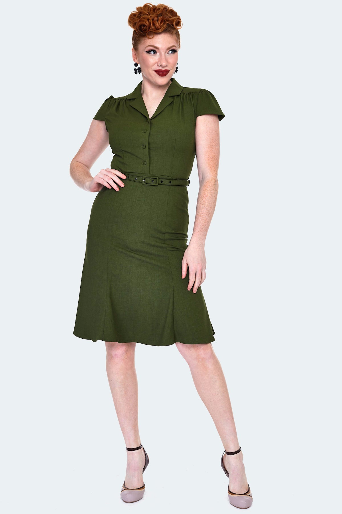 A model wearing a dark olive green shirtwaist dress with a v-neck and notched collar. It has gathering at the shoulders and slightly puffed cap sleeves. Hitting at the natural waist with a self belt and ends just above the knee with a slight flare. Seen from the front