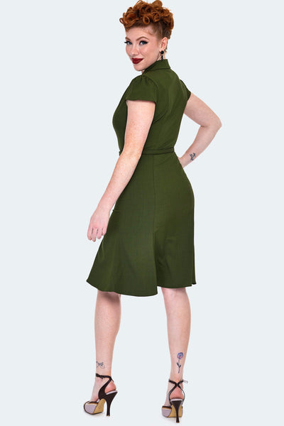 A model wearing a dark olive green shirtwaist dress with a v-neck and notched collar. It has gathering at the shoulders and slightly puffed cap sleeves. Hitting at the natural waist with a self belt and ends just above the knee with a slight flare. Seen from the back
