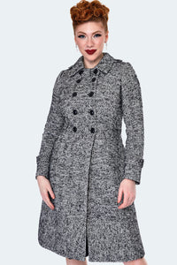 Model wearing a black and white herringbone pattern double breasted pea coat with black buttons down the front and at each cuff, faux epaulets, a wide waistband, and fit and flare shape ending just at the knee. Shown buttoned up from the front 