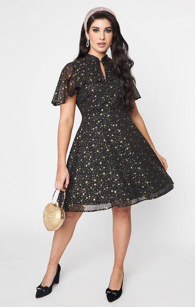 A black crepe chiffon dress with an all-over pattern of small gold stars and starbursts. It has a mandarin collar with keyhole and an attached caplet with a ruffled hem. The full skirt ends just above the knee. Shown from the front worn by a model 