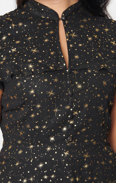 A black crepe chiffon dress with an all-over pattern of small gold stars and starbursts. It has a mandarin collar with keyhole and an attached caplet with a ruffled hem. The full skirt ends just above the knee. Shown from the front worn by a model in close up