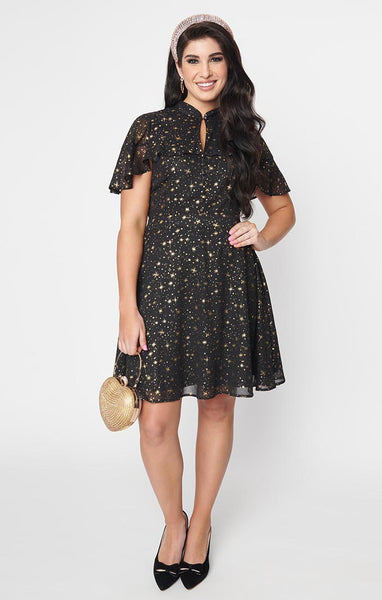 A black crepe chiffon dress with an all-over pattern of small gold stars and starbursts. It has a mandarin collar with keyhole and an attached caplet with a ruffled hem. The full skirt ends just above the knee. Shown from the front worn by a model