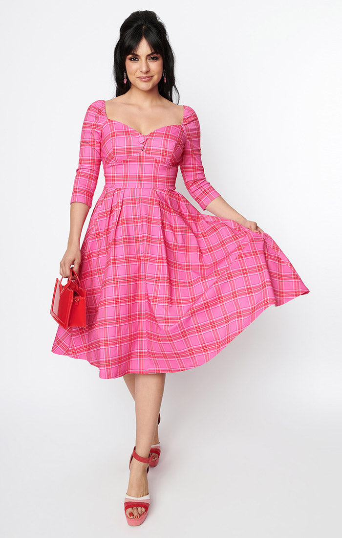 Model wearing a swing dress in a pink, red, and white plaid pattern with 3/4 sleeves, sweetheart neckline with button detail, open square back, and a full knee length skirt. Shown from the front
