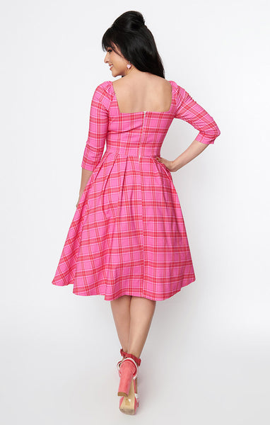 Model wearing a swing dress in a pink, red, and white plaid pattern with 3/4 sleeves, sweetheart neckline with button detail, open square back, and a full knee length skirt. Shown from the back