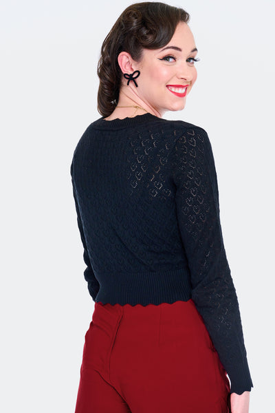 Model wearing black v-neck cardigan with openwork heart design, scalloped edges, and black plastic heart-shaped buttons. Shown from the back buttoned up