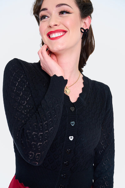 Model wearing black v-neck cardigan with openwork heart design, scalloped edges, and black plastic heart-shaped buttons. Shown from the front buttoned in closeup 