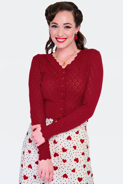Model wearing deep red  v-neck cardigan with openwork heart design, scalloped edges, and black plastic heart-shaped buttons. Shown from the front buttoned up
