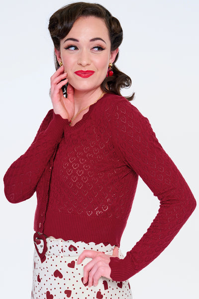 Model wearing deep red v-neck cardigan with openwork heart design, scalloped edges, and black plastic heart-shaped buttons. Shown from the side buttoned up