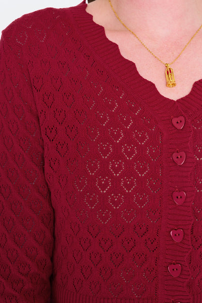 Model wearing deep red v-neck cardigan with openwork heart design, scalloped edges, and black plastic heart-shaped buttons. Shown from the front buttoned up in closeup