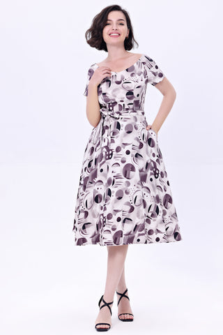 Model wearing a cocktail length short sleeved dress with a full gathered skirt and surplice style v neckline. It has a white background patterned with achromatic grey and black abstract shapes. Shown from front