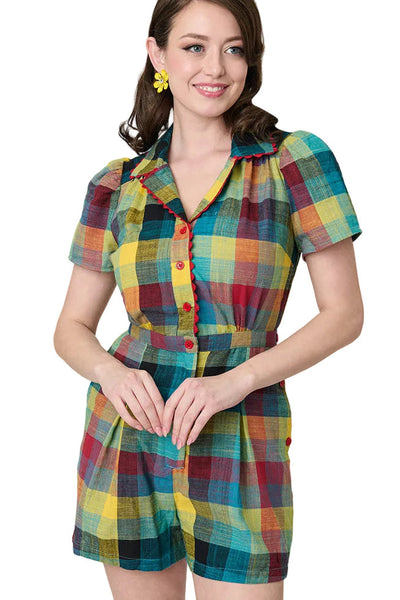 Model wearing cotton red, blue, and yellow Madras plaid print romper with pointed collar with red rickrack trim and red plastic button-up bodice, puffed short sleeves, and pleated relaxed fit shorts with side seam pockets and matching buttons down side of left hip. Shown from front in close up