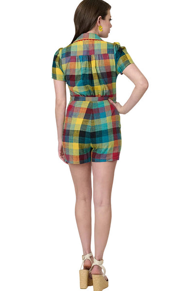 Model wearing cotton red, blue, and yellow Madras plaid print romper with pointed collar with red rickrack trim and red plastic button-up bodice, puffed short sleeves, and pleated relaxed fit shorts with side seam pockets and matching buttons down side of left hip. Shown from back