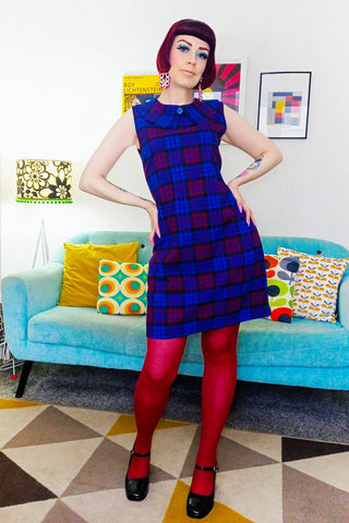 blue, berry, and bblack plaid 60s inspired a-line sleeveless shift dress is in a mid-thigh mini length and features a Buster Brown collar finished with a decorative blue floral button at the neckline. Shown on a model.