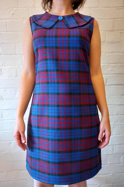 blue, berry, and bblack plaid 60s inspired a-line sleeveless shift dress is in a mid-thigh mini length and features a Buster Brown collar finished with a decorative blue floral button at the neckline. Shown on a model.