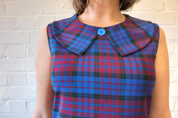 blue, berry, and bblack plaid 60s inspired a-line sleeveless shift dress is in a mid-thigh mini length and features a Buster Brown collar finished with a decorative blue floral button at the neckline. Showing close up of collare, on a model.