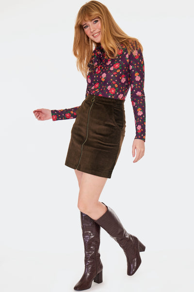 a model wearing a brown corduroy high-waisted mini skirt with side seam pockets and front zip closure with brass tone o-ring zipper pull. Shown from the front