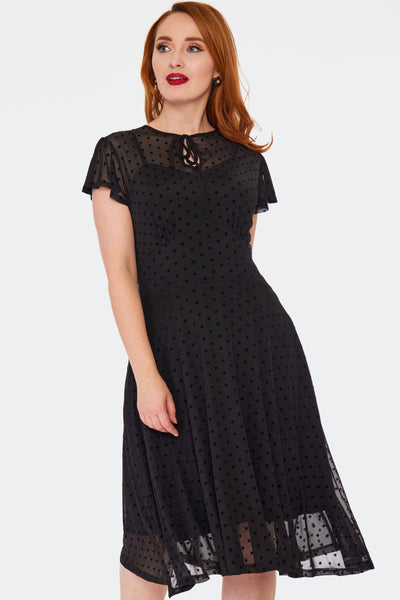 a model wearing a flutter sleeve flare dress made of black flocked polka dot mesh. It has tie keyhole detail at its jewel neckline, gathered underbust seaming, a below the knee flared skirt, and an attached black slip. Shown from the front