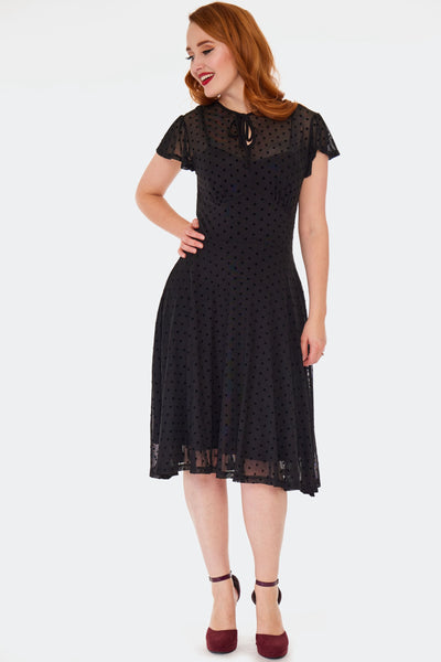 a model wearing a flutter sleeve flare dress made of black flocked polka dot mesh. It has tie keyhole detail at its jewel neckline, gathered underbust seaming, a below the knee flared skirt, and an attached black slip. Shown from the front